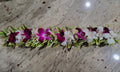 A floral lei or flower garland laid horizontally on a speckled beige marble surface, featuring white and deep magenta orchids with green and yellow variegated leaves.