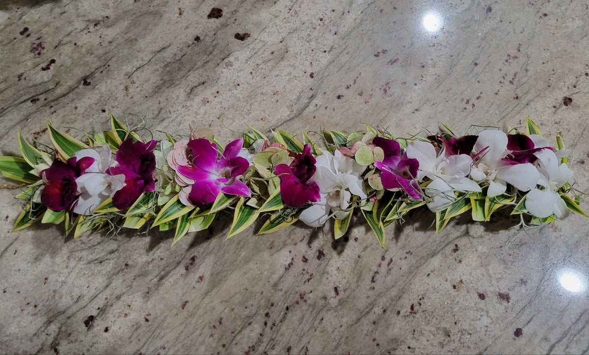 A floral lei or flower garland laid horizontally on a speckled beige marble surface, featuring white and deep magenta orchids with green and yellow variegated leaves.